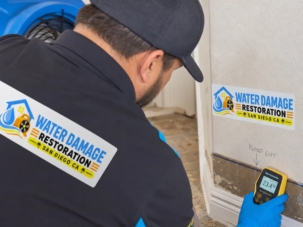 Moisture meter reading on drywall near a marked baseboard flood-cut line with drying equipment in the background